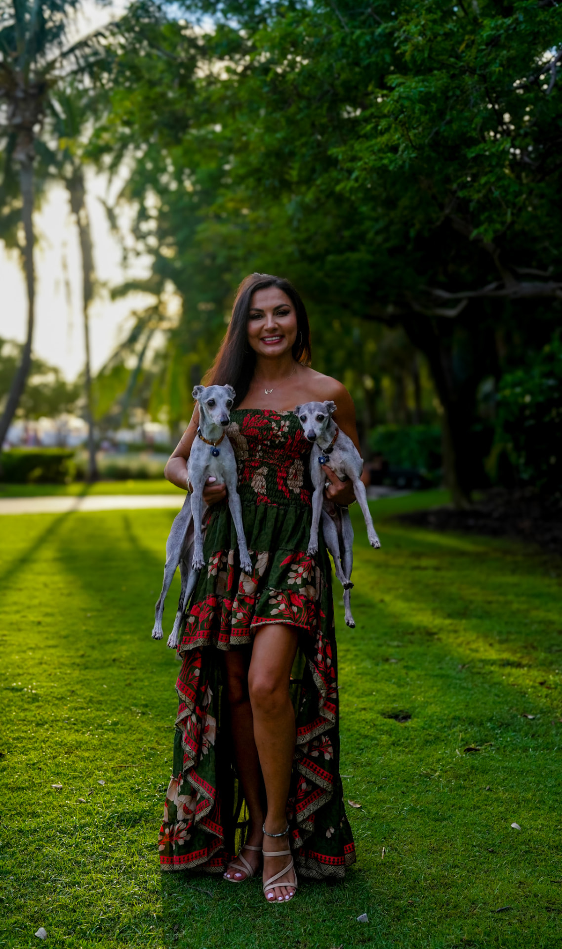 Elmira carrying both greyhounds along a palm-lined lawn at golden hour