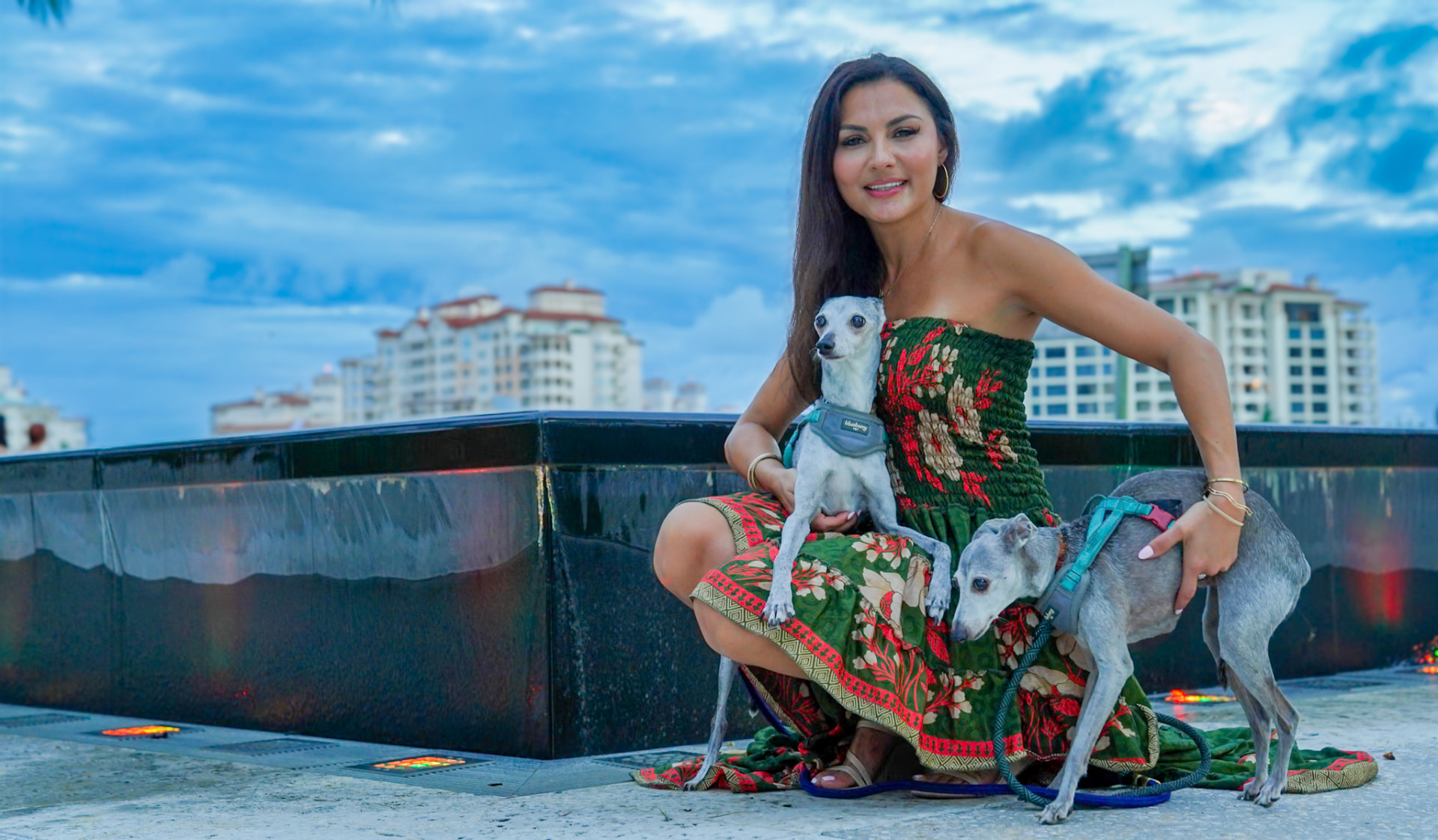 Elmira holding one greyhound close while her hand rests on the other, water and sky behind them